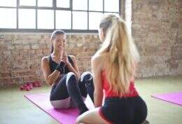 Duas mulheres em ambiente de academia. Uma sorri para a foto enquanto executa abdominais. A segunda, de costas para a foto, parece estar instruindo a primeira.