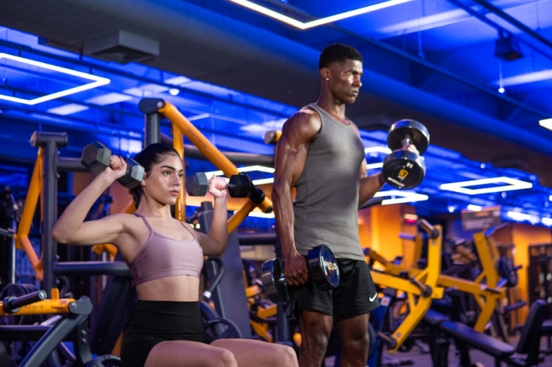 Imagem de um homem e uma mulher treinando musculação, com o homem em pé realizando exercício para os músculos superiores e a mulher sentada, ambos focados no treino para ganho de massa muscular.