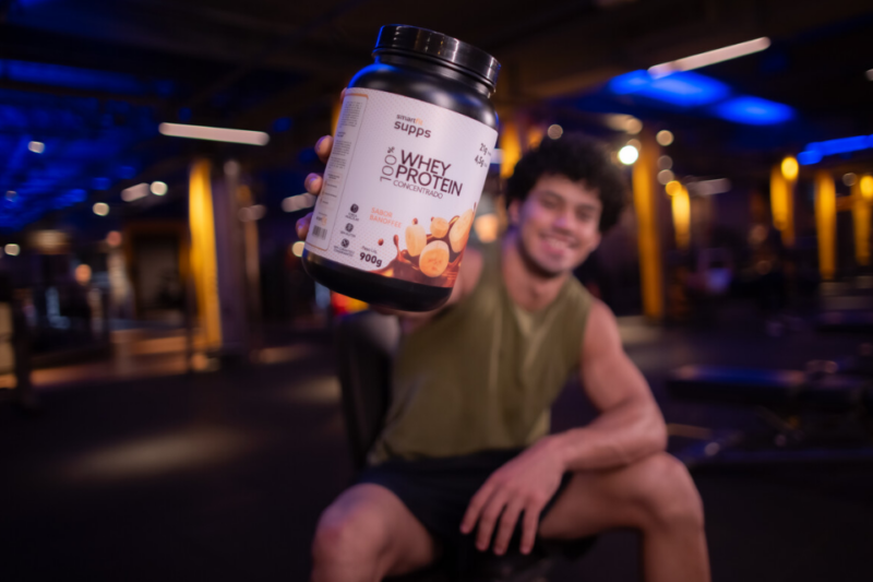 A imagem apresenta um homem em uma unidade da Smart Fit apresentando o Whey do Smart Fit Supps