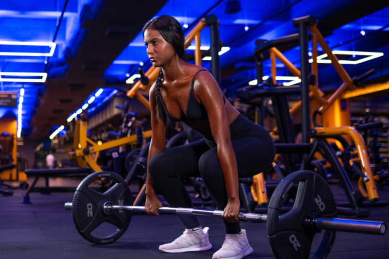 A imagem apresenta uma mulher concentrada em uma unidade da Smart Fit se preparando para realizar um exercício com a barra.