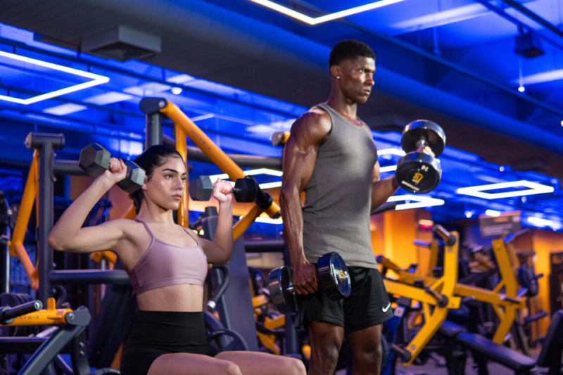 A imagem apresenta um homem e uma mulher em uma unidade da Smart Fit. Ela está sentada fazendo um exercício para ombros, enquanto ele está em pé trabalhando os biceps.