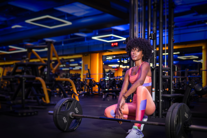 A imagem apresenta uma mulher agachada próxima a uma barra na Smart FIt, com cerca de 20 kg de cada lado na barra.