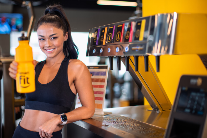 A imagem apresenta uma mulher em uma unidade da Smart Fit bebendo uma bebida na garrafinha da marca ao lado de um totem de bebidas